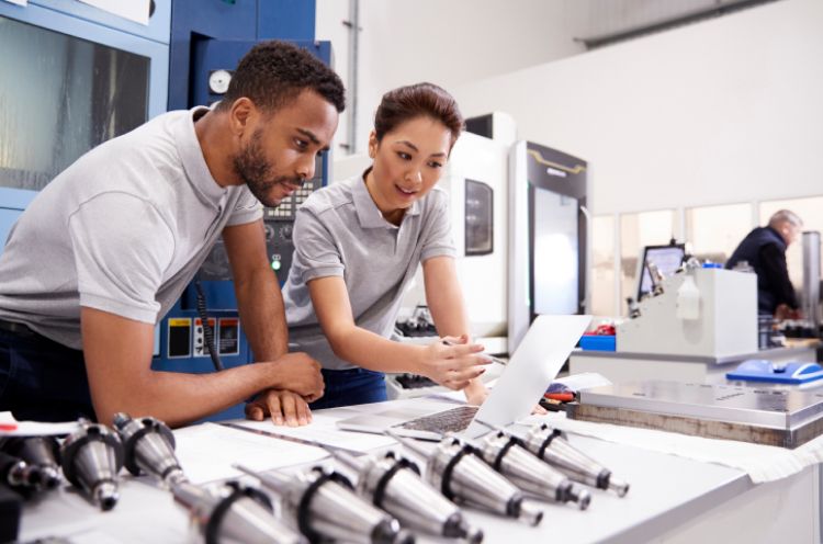 Two engineers in gray polos review data on a laptop while collaborating in an industrial setting. They are surrounded by various engineering tools and components, highlighting teamwork in civil engineering and system integration.