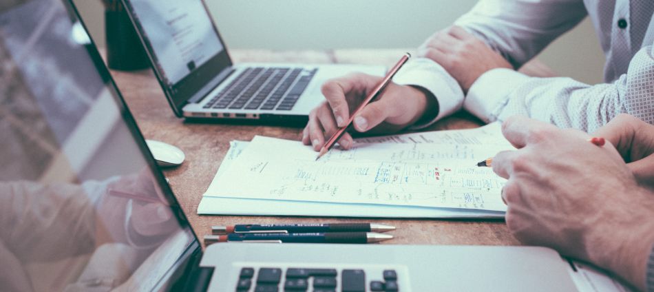 Two individuals are collaborating at a desk, focused on project documents. Laptops are open nearby, and one person is writing on the papers while the other points to the notes, highlighting teamwork in engineering discussions.