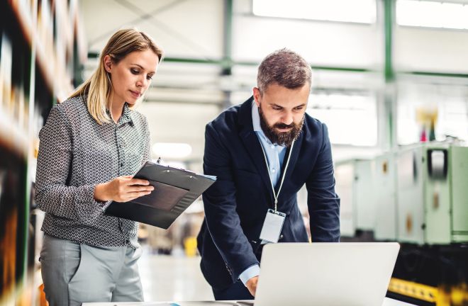 Two professionals are collaborating in an industrial setting. A woman holds a clipboard while a man reviews information on a laptop. They are engaged in discussion, likely focused on engineering or project management tasks related to structural engineering.