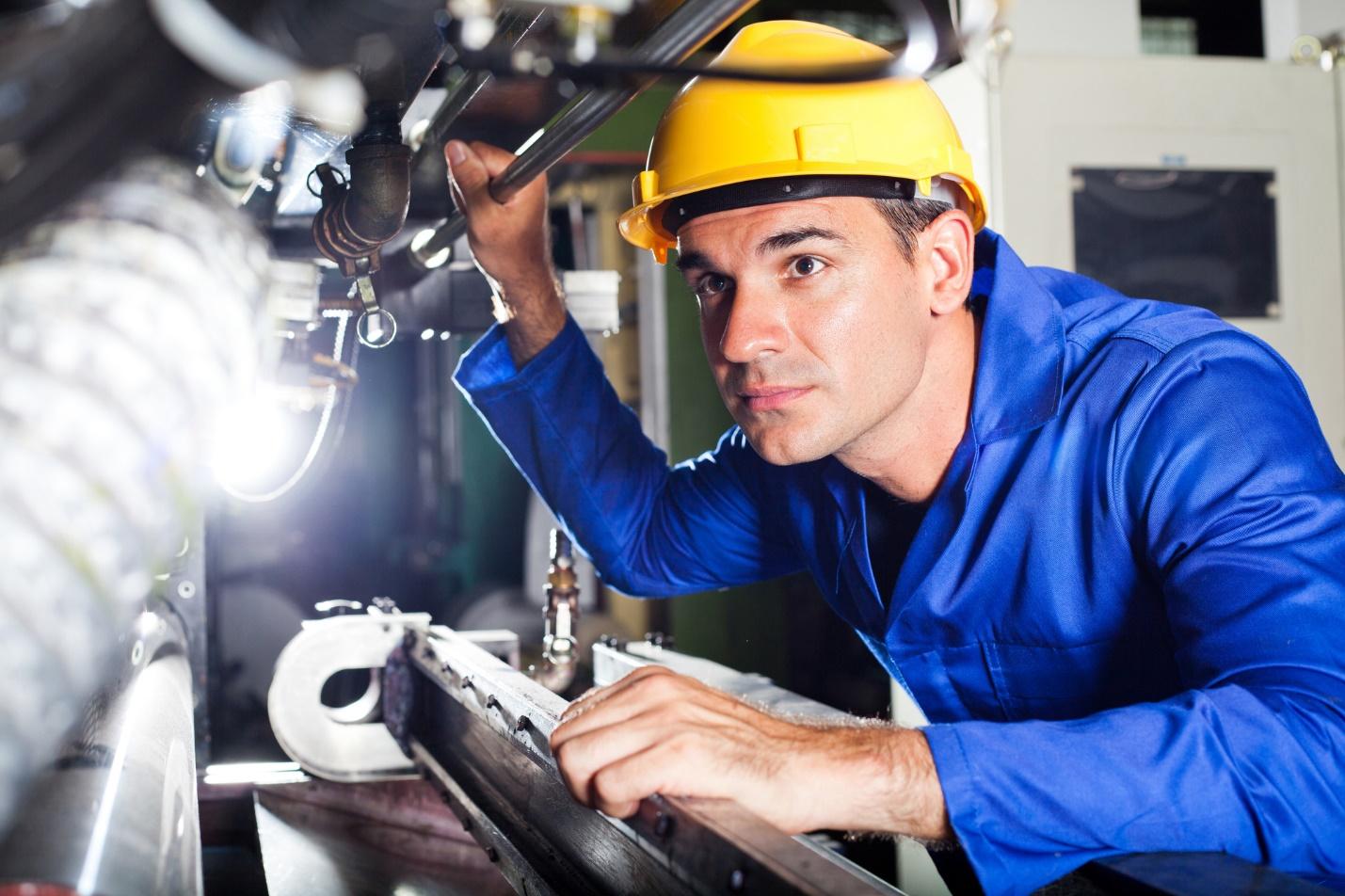 A focused engineer in a blue work uniform and a yellow hard hat inspects industrial equipment. He is closely examining machinery components in a pilot plant, demonstrating hands-on involvement in engineering tasks for system integration and digital transformation.