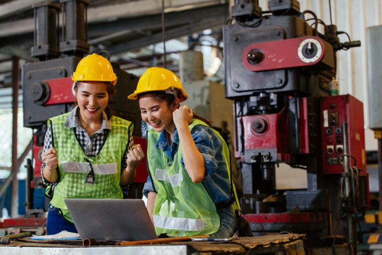 Two mechanical engineers in safety gear celebrating successful coordination in a factory setting, standing in front of industrial machinery and a laptop.