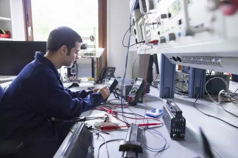 A technician performing intricate, close-up electronic testing on a control system component or instrument at a workbench with specialized test equipment like multimeters.