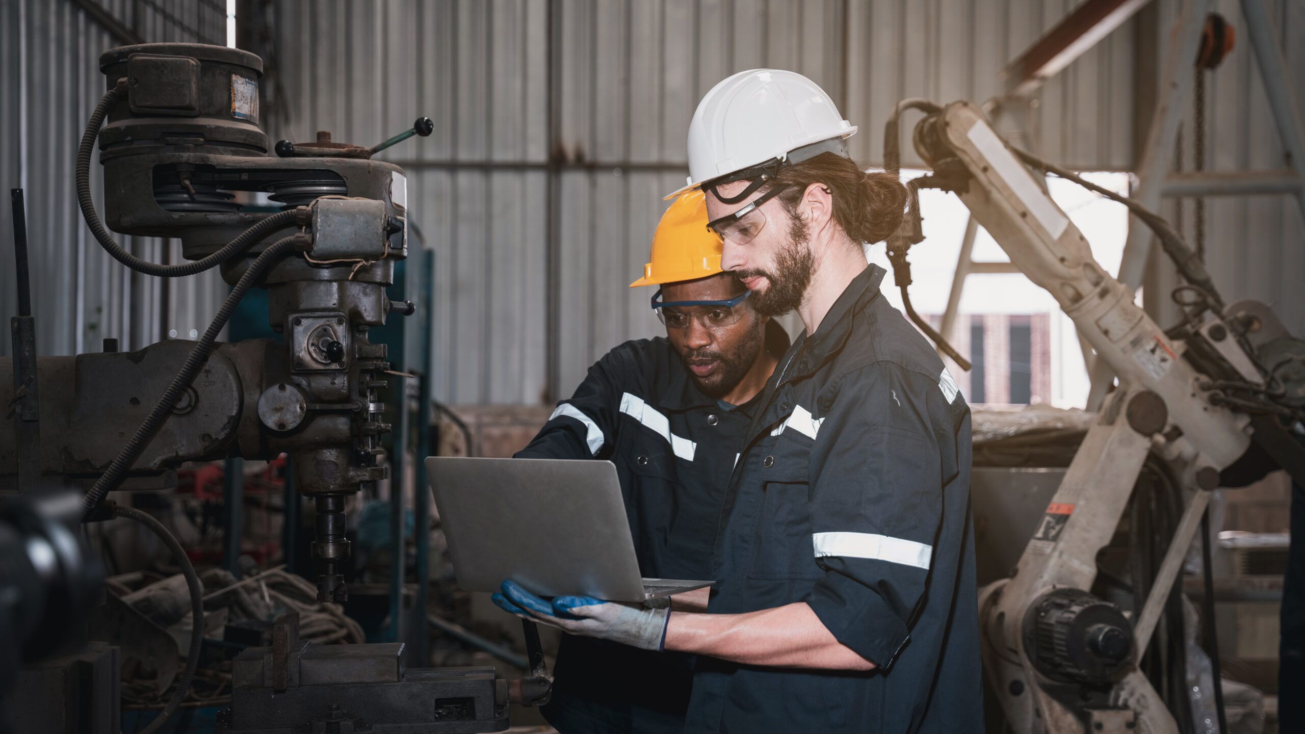 Industrial engineers reviewing maintenance data on a laptop beside machinery in a manufacturing facility